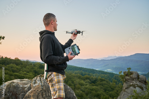 Man operating drone using remote controller. Man using drone at sunset for photos and video making while standing on top of high boulder in the mountains.