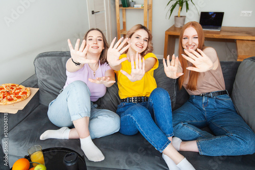 three girls at home are waving their hands. students are sitting on the couch with pizza
