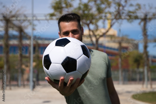 Handsome young man holds a football in his hand and shows it to the camera in focus. The man is a sports fan and in particular a football fan. He is a fan of his team. Concept of sports and team play. © @skuder_photographer