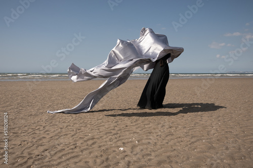 art portrait of woman in long black dress on the beach waving silver fabric photo