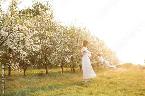 Mom holds her son's arms and spins around. Blooming garden in spring. © Yuliia