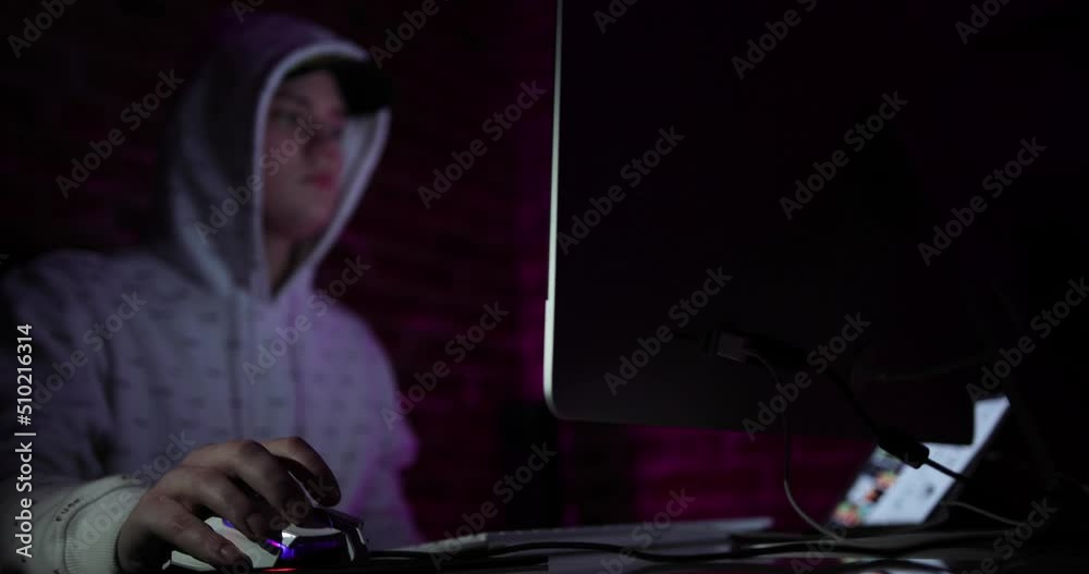 attractive young man in cap and hood working at computer in dark room ...