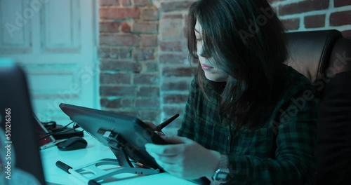 attractive hipster girl with a piercing in the nose works at the tablet. girl artist draws on the tablet. girl sitting in a chair in an office loft style. behind a brick wall and a white shabby door. photo