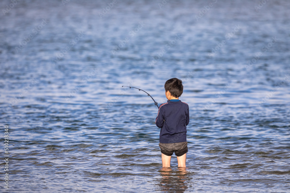 A young unidentifiable boy with a fishing rod stands in a shallow lake, surrounded by calm blue water, on a sunny afternoon. Copy space.