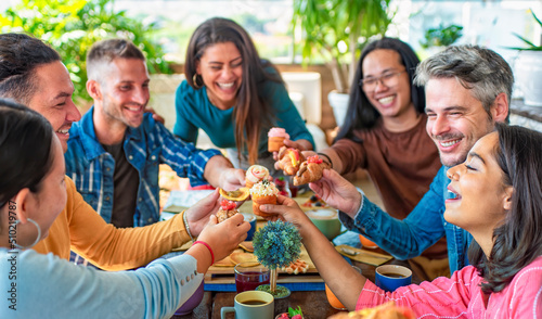 multiethnic large group of friends sitting on cafe table restaurant eating a muffin making faces. diverse people celerating sweet breakfast together enjoying happy holidays. lifestyle and joy concept