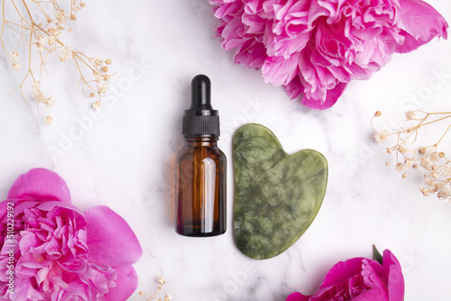 Glass bottle with oil, jade Gua sha stone for face massage on marble background with peony flowers