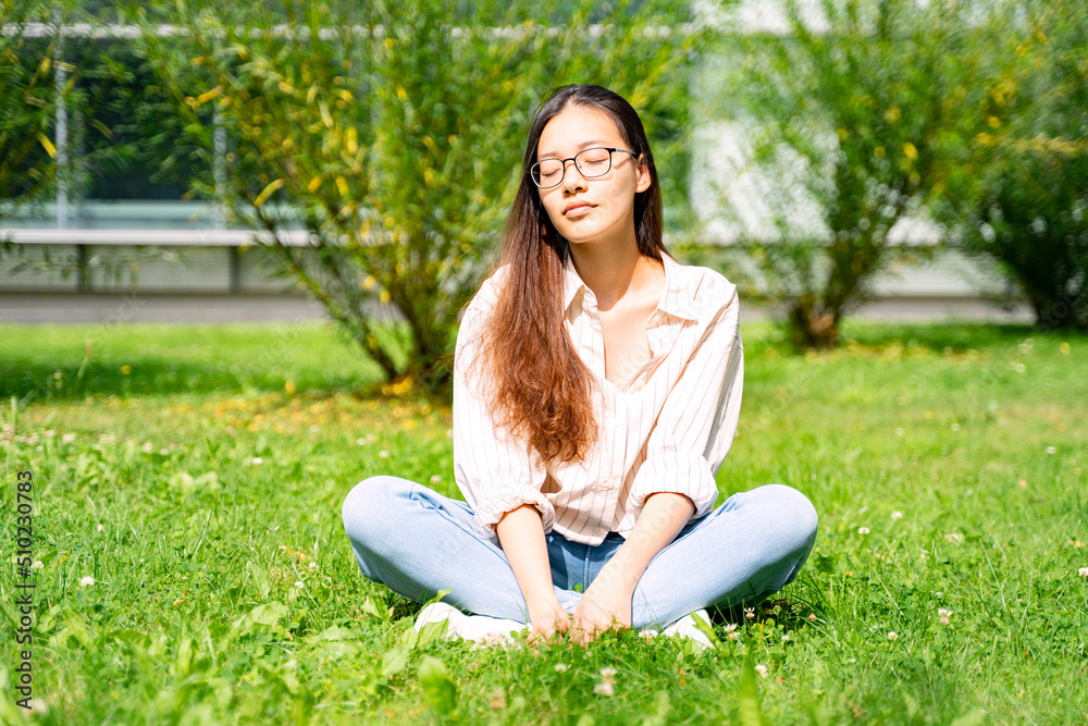 Young Asian Thai, Vietnamese or Chinese woman in shirt, jeans and glasses sitting relaxing and dreaming on green grass loan near bushes and office building on weekend or holiday on summer sunny day. 