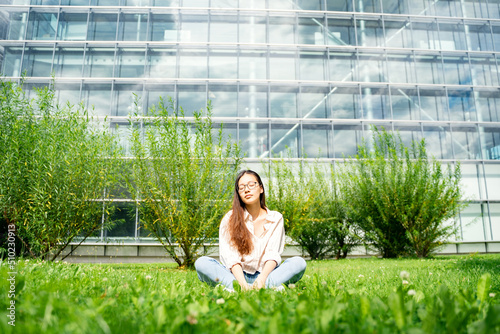 Photography Young Asian Thai, Vietnamese or Chinese woman in casual clothes and glasses sitting relaxing and dreaming on grass loan near bushes and glass office building on weekend after work on summer day