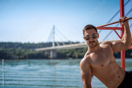 Shirtless muscular man in sunglasses on a beach.