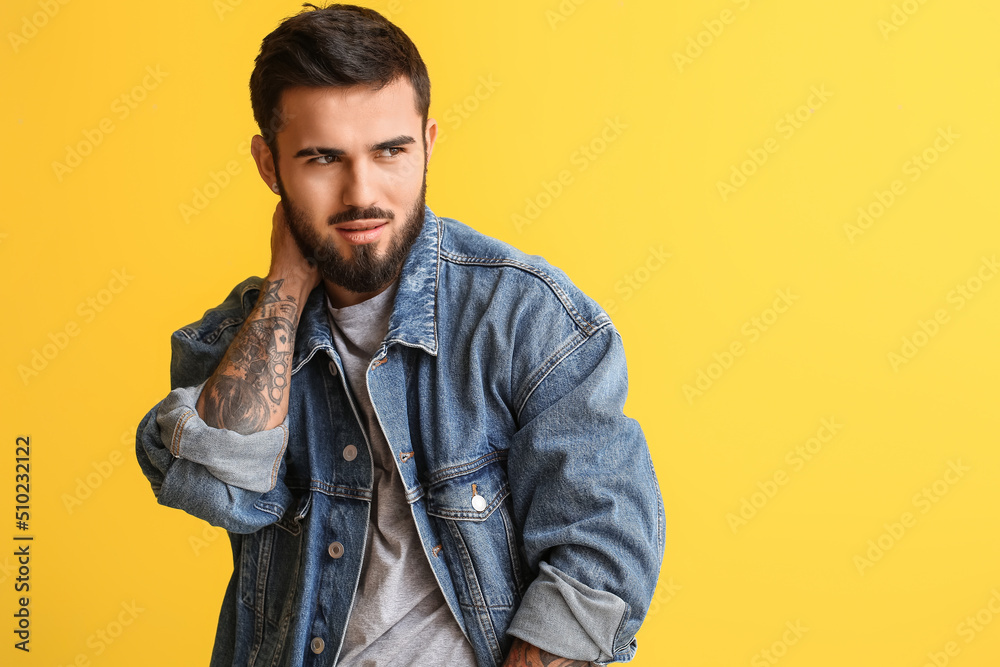 Fototapeta premium Stylish young man with beard and tattoos wearing denim jacket posing confidently on yellow background, he looks to the side with a thoughtful expression, his modern fashion sense