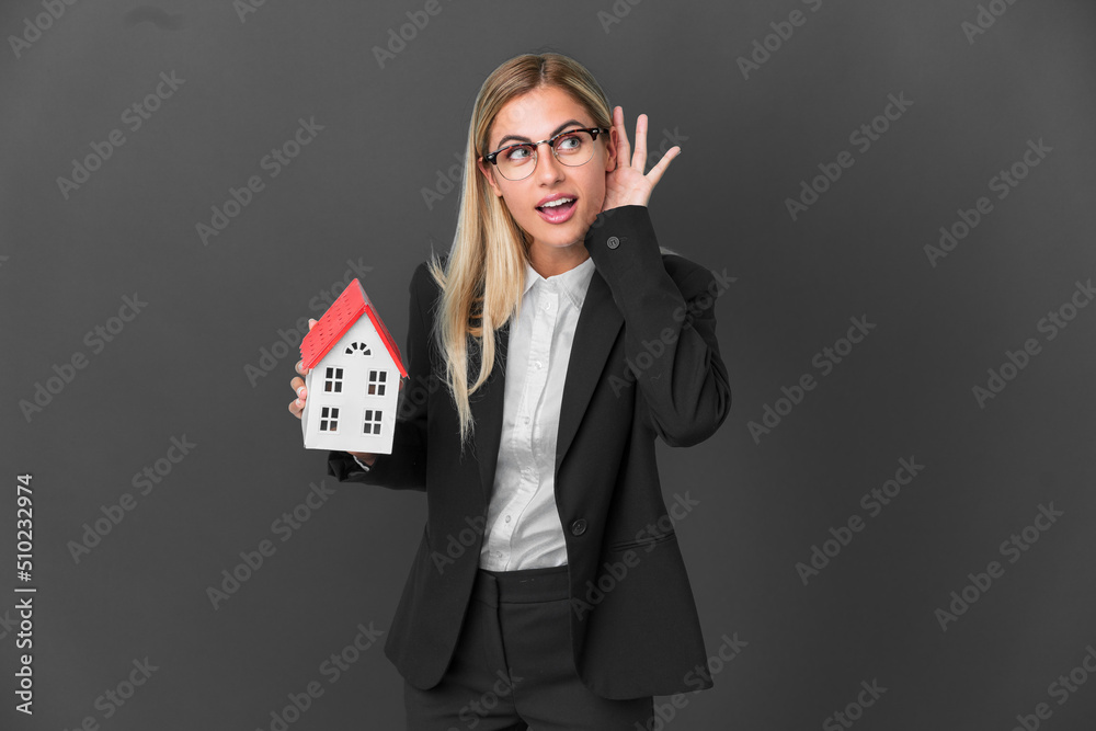 Blonde Uruguayan girl holding a house toy isolated on black background listening to something by putting hand on the ear Blonde Uruguayan girl holding a house toy isolated on black background listening to something by putting hand on the ear