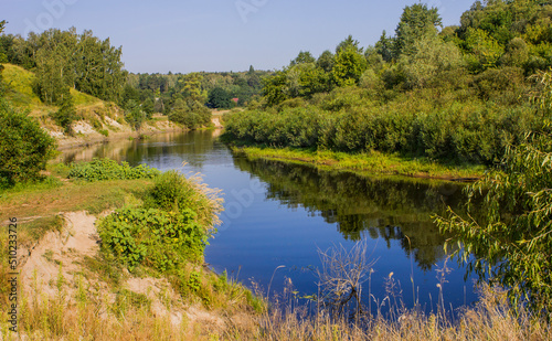a river with green steep banks and the reflection of trees in the water on a summer sunny day