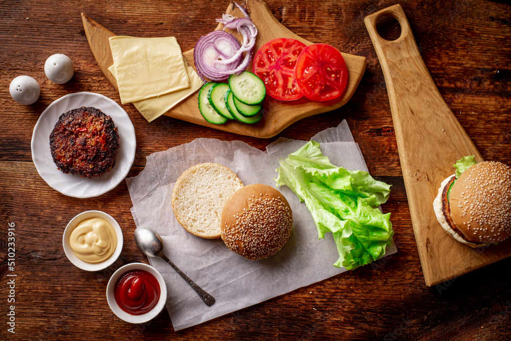 Ingredients for a classic hamburger. Grilled meat, vegetables, greens, sauces near a sesame bun. Wooden background. Cooking hamburger or cheeseburger. Ingredients for a classic hamburger. Grilled meat, vegetables, greens, sauces near a sesame bun. Wooden background. Cooking hamburger or cheeseburger.
