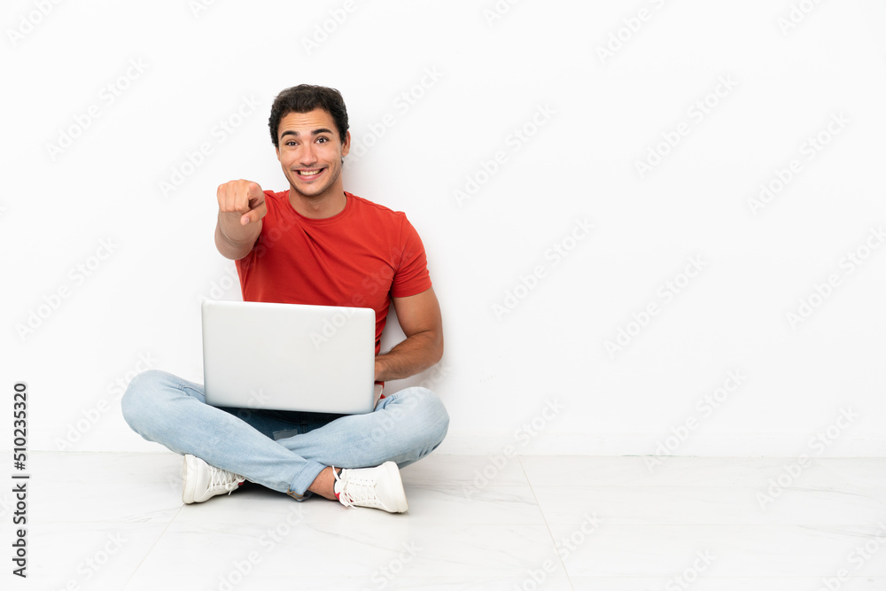 Caucasian handsome man with a laptop sitting on the floor points finger at you with a confident expression Caucasian handsome man with a laptop sitting on the floor points finger at you with a confident expression