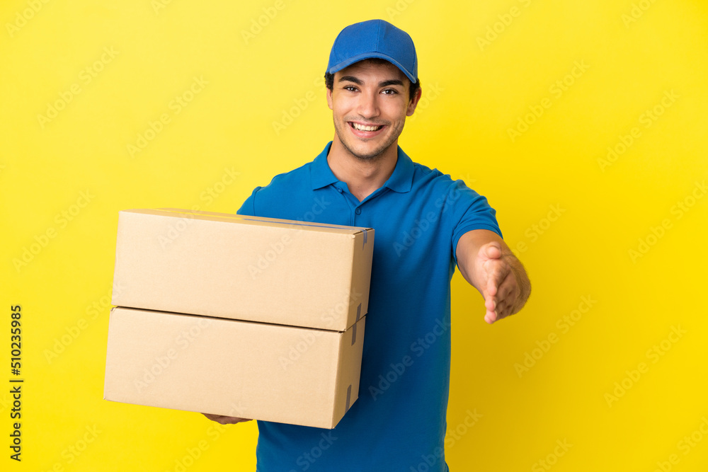 Delivery man over isolated yellow wall shaking hands for closing a good deal Delivery man over isolated yellow wall shaking hands for closing a good deal