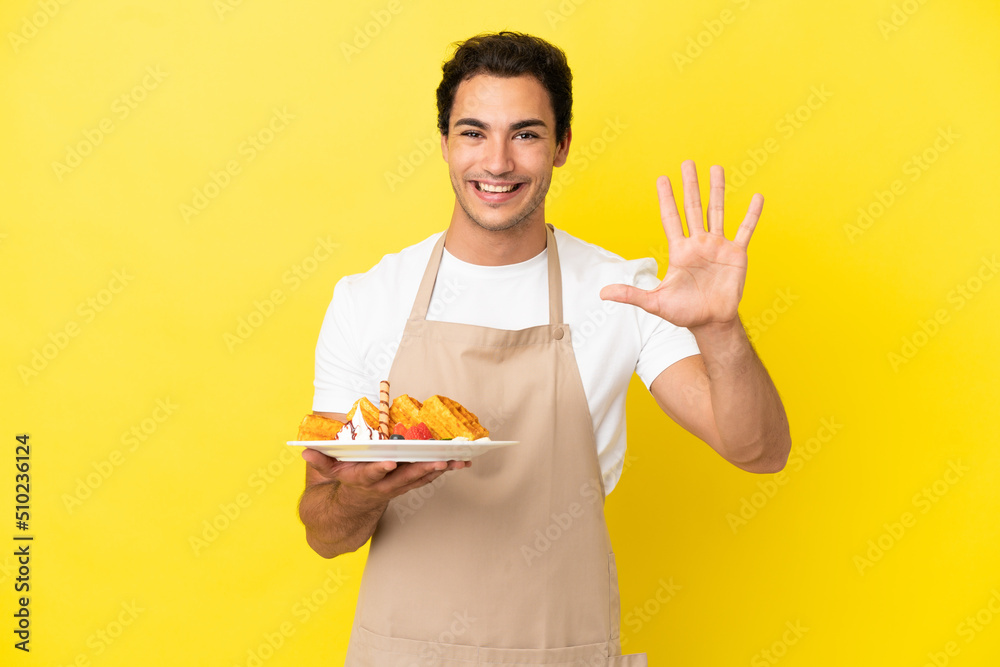 Restaurant waiter holding waffles over isolated yellow background counting five with fingers Restaurant waiter holding waffles over isolated yellow background counting five with fingers