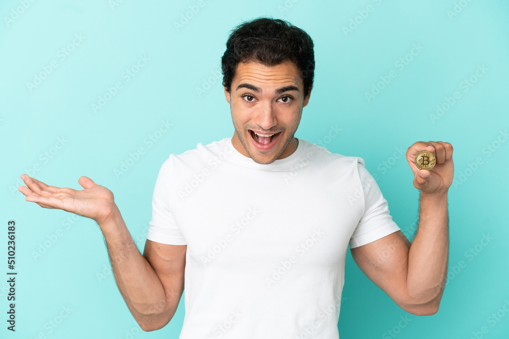 Young man holding a Bitcoin over isolated blue background with shocked facial expression Young man holding a Bitcoin over isolated blue background with shocked facial expression