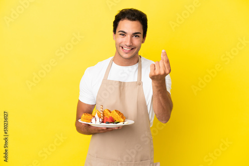 Restaurant waiter holding waffles over isolated yellow background making money gesture
