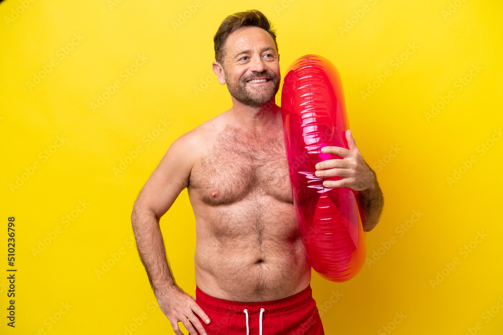 Middle age caucasian man holding inflatable donut isolated on yellow background posing with arms at hip and smiling