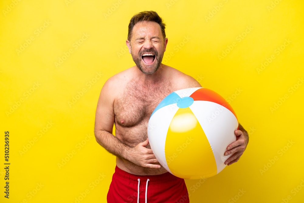 Middle age caucasian man holding beach ball isolated on yellow background smiling a lot