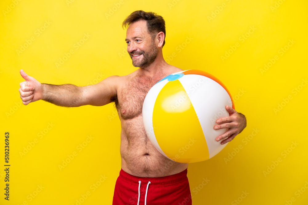 Middle age caucasian man holding beach ball isolated on yellow background giving a thumbs up gesture