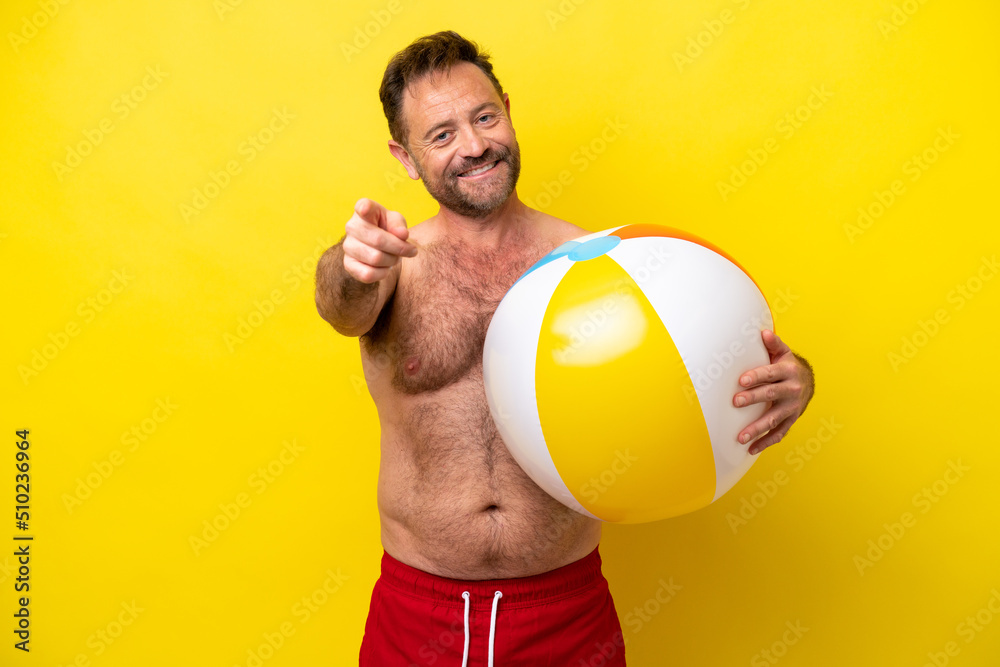 Middle age caucasian man holding beach ball isolated on yellow background pointing front with happy expression Middle age caucasian man holding beach ball isolated on yellow background pointing front with happy expression