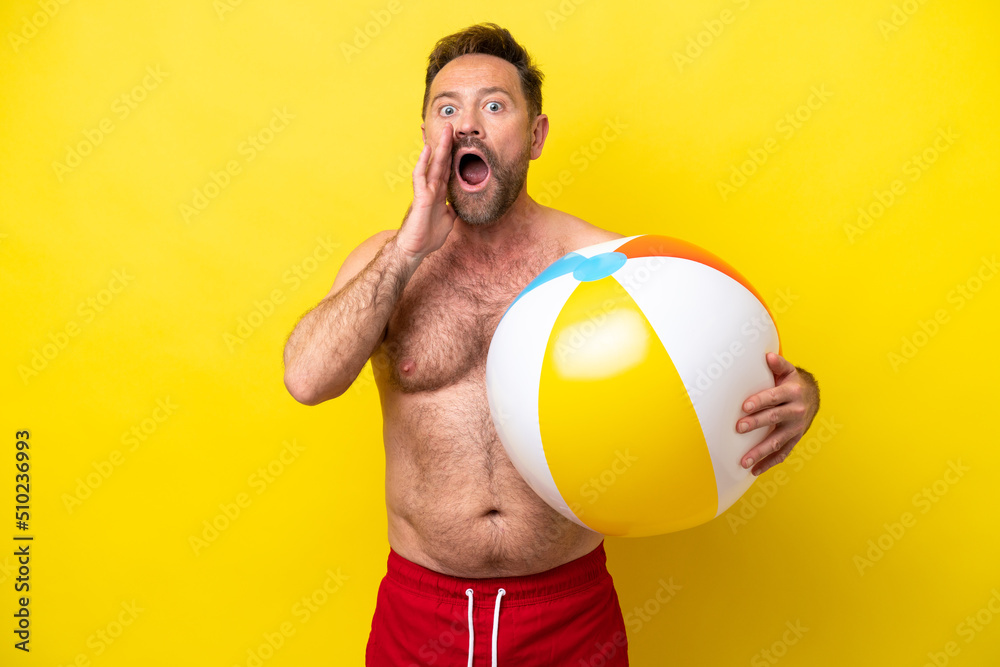 Middle age caucasian man holding beach ball isolated on yellow background with surprise and shocked facial expression