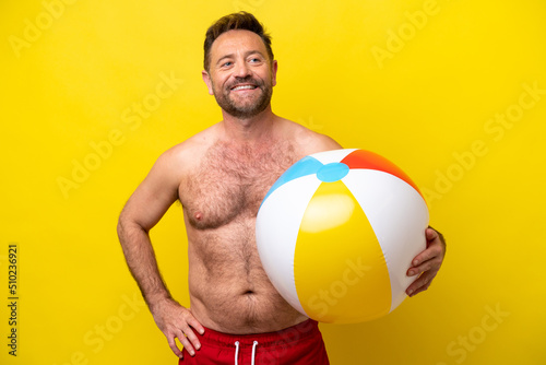 Middle age caucasian man holding beach ball isolated on yellow background posing with arms at hip and smiling