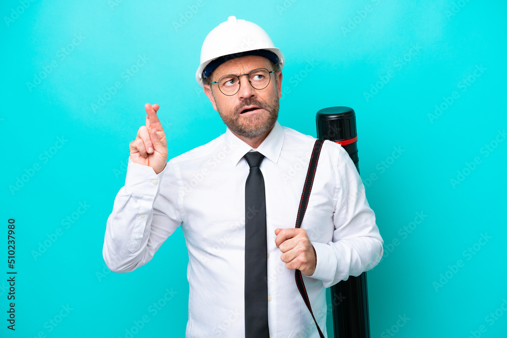 Middle age architect man with helmet and holding blueprints isolated on blue background with fingers crossing and wishing the best Middle age architect man with helmet and holding blueprints isolated on blue background with fingers crossing and wishing the best