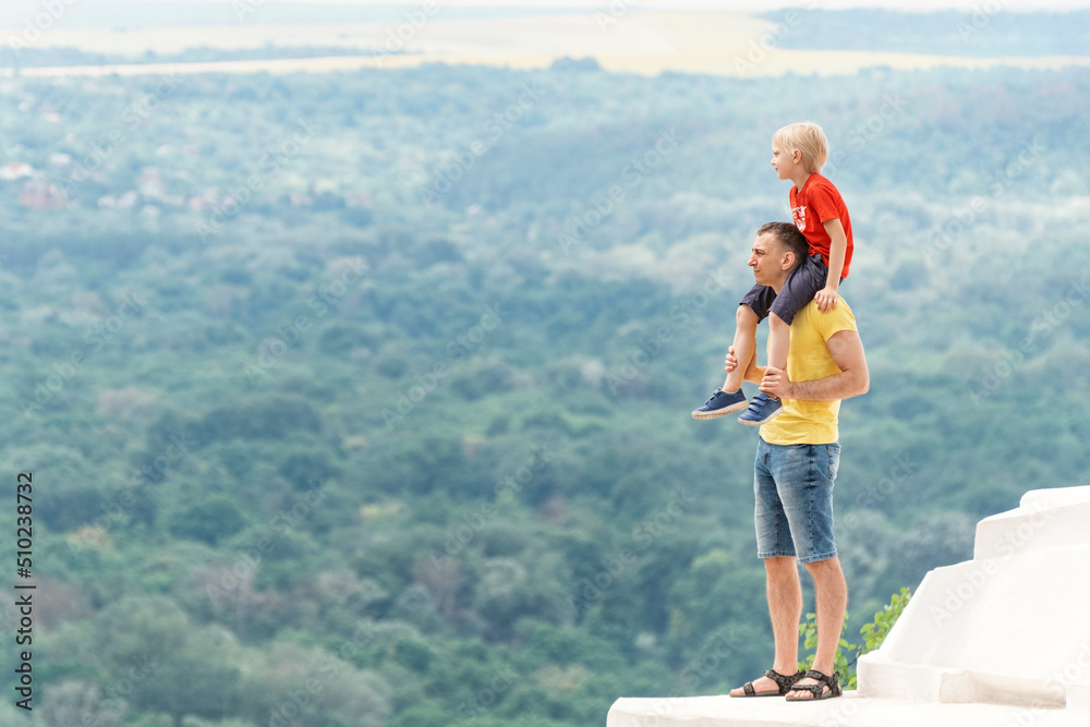 Son sits on his fathers shoulders on forest background. Full-length portrait of dad and son on the observation deck.