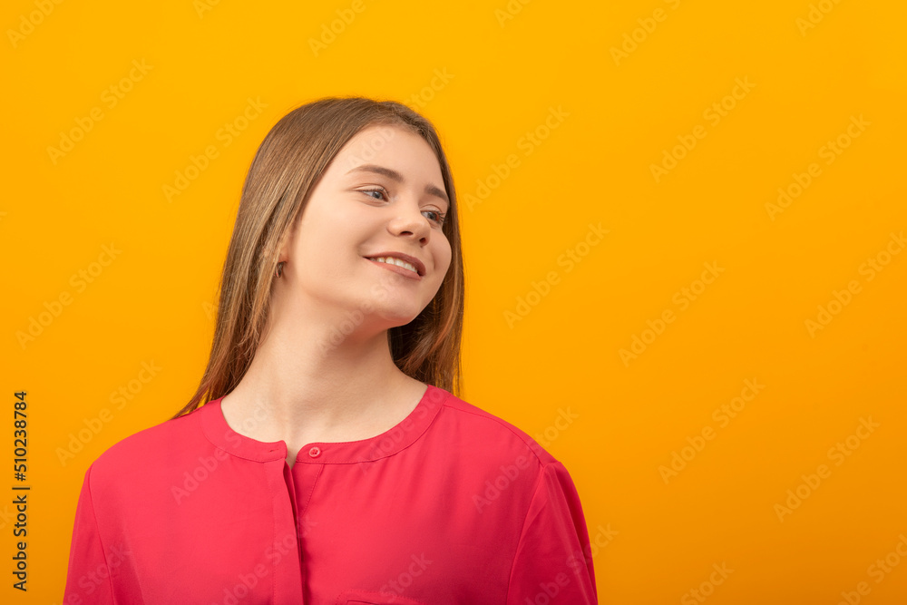 Fototapeta premium Close up portrait of young girl smiling sweetly and looking away. Teenage girl in red shirt on an orange background.