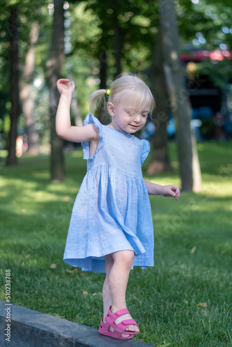 Portrait of little blonde girl in blue dress in park. Three-year-old child in sundress with two tails on trees background.
