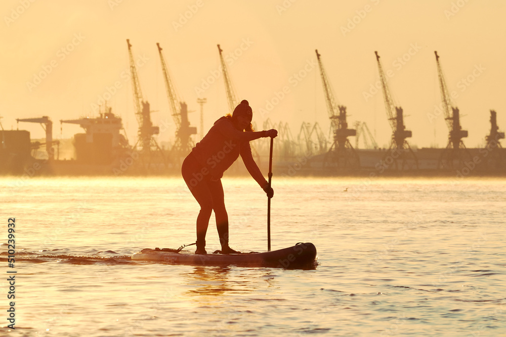Silhouette of middle-aged woman stand up paddle boards on a Danube river at winter sunrise against the backdrop of port cranes