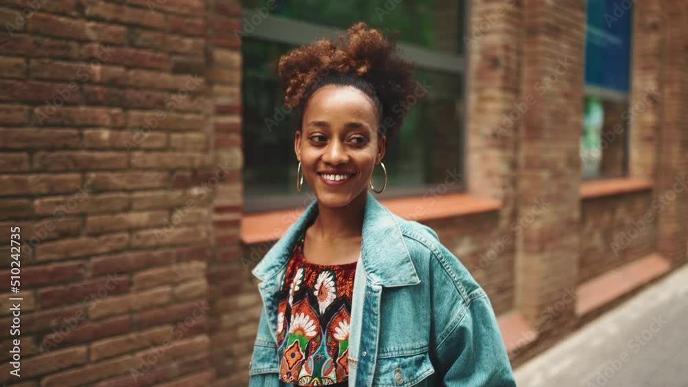 Close-up African girl with ponytail wearing denim jacket, in crop top with national pattern walking smiling down the street against modern buildings background. Slow motion Close-up African girl with ponytail wearing denim jacket, in crop top with national pattern walking smiling down the street against modern buildings background. Slow motion