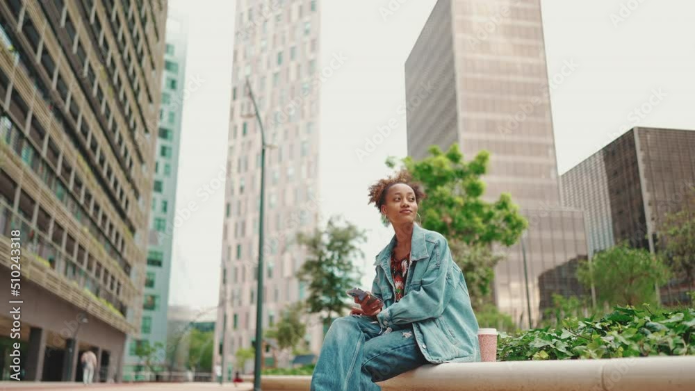 African girl with ponytail, wearing denim jacket, in crop top with national pattern with disposable cup of coffee sits with mobile phone in her hands and turns her head against buildings background African girl with ponytail, wearing denim jacket, in crop top with national pattern with disposable cup of coffee sits with mobile phone in her hands and turns her head against buildings background