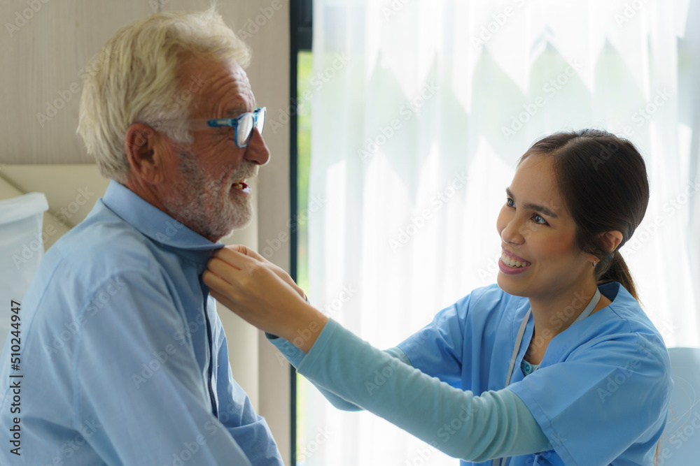 Obraz premium Young Asian woman nurse helping get dress to disabled elderly man in bed at retirement home. Millennial caregiver assisting handicapped senior patient, taking care of older male indoors