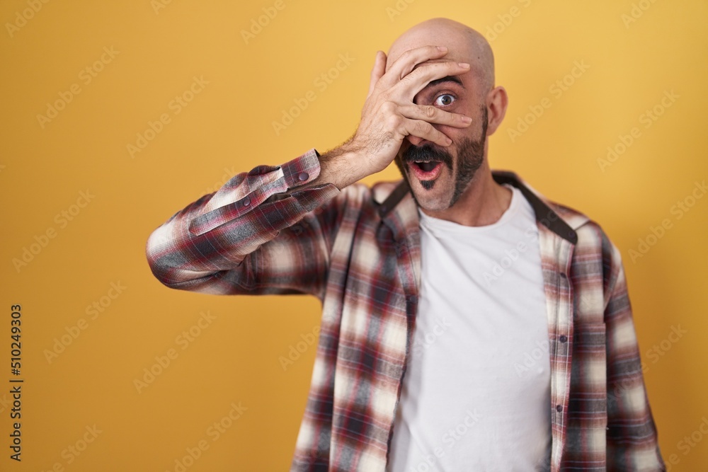 Hispanic man with beard standing over yellow background peeking in shock covering face and eyes with hand, looking through fingers with embarrassed expression. Hispanic man with beard standing over yellow background peeking in shock covering face and eyes with hand, looking through fingers with embarrassed expression.