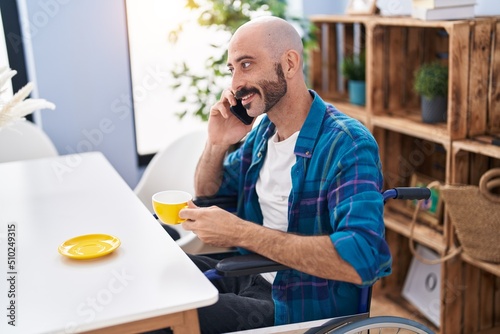 Young hispanic man sitting on wheelchair talking on smartphone drinking coffee at home