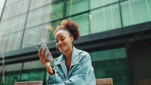 Cute African girl with ponytail, wearing denim jacket, in crop top with national pattern, sitting on bench and using smartphone on modern buildings background. Slow motion