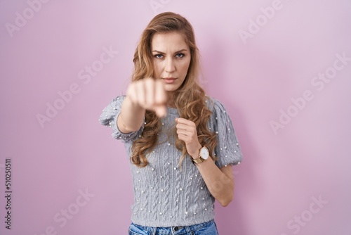 Beautiful blonde woman standing over pink background punching fist to fight, aggressive and angry attack, threat and violence