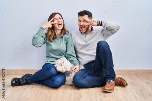 Young hispanic couple sitting on the floor with dog doing peace symbol with fingers over face, smiling cheerful showing victory