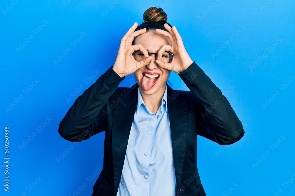 Young hispanic girl wearing business clothes and glasses doing ok gesture like binoculars sticking tongue out, eyes looking through fingers. crazy expression.
