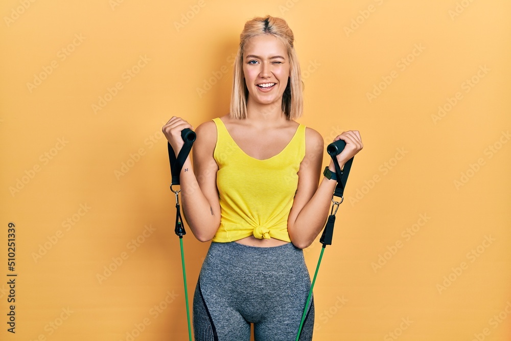 Beautiful blonde woman training arm resistance with elastic arm bands winking looking at the camera with sexy expression, cheerful and happy face.