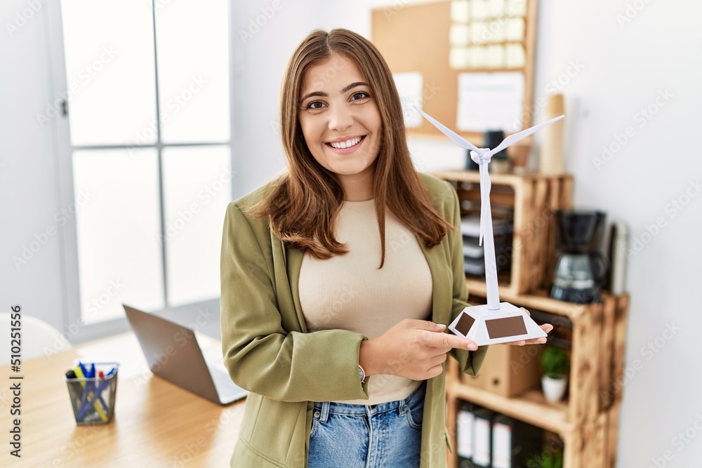 Young brunette woman holding solar windmill for renewable electricity at the office looking positive and happy standing and smiling with a confident smile showing teeth