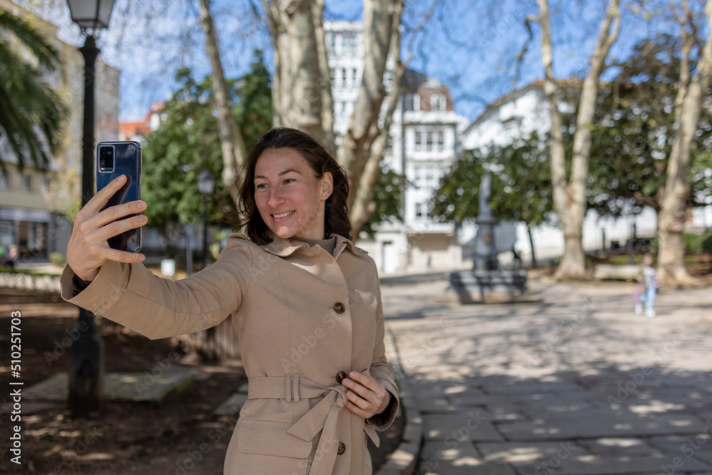 Obraz premium A girl in a beige raincoat stands in the park and waves her hand at the phone. The girl is smiling, she is happy. It's beautiful sunny outside, blue clear skies. The smell of freedom.