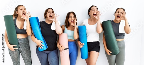 Group of women holding yoga mat standing over isolated background shouting and screaming loud to side with hand on mouth. communication concept.
