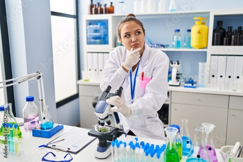 Young hispanic woman working at scientist laboratory serious face thinking about question with hand on chin, thoughtful about confusing idea