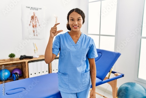 Beautiful hispanic physiotherapist woman at pain recovery clinic smiling and confident gesturing with hand doing small size sign with fingers looking and the camera. measure concept.