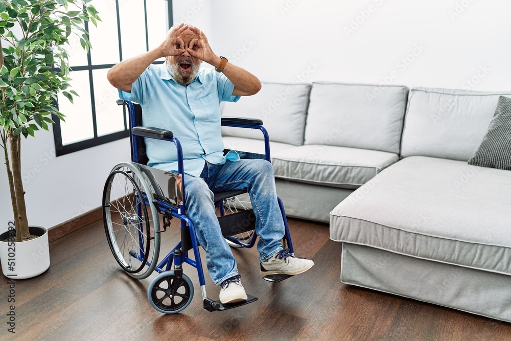 Handsome senior man sitting on wheelchair at the living room doing ok gesture like binoculars sticking tongue out, eyes looking through fingers. crazy expression. Handsome senior man sitting on wheelchair at the living room doing ok gesture like binoculars sticking tongue out, eyes looking through fingers. crazy expression.