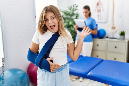 Blonde little girl wearing arm on sling at rehabilitation clinic celebrating victory with happy smile and winner expression with raised hands